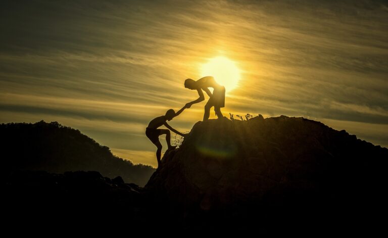 Photograph of sunset with one person helping another up a hill